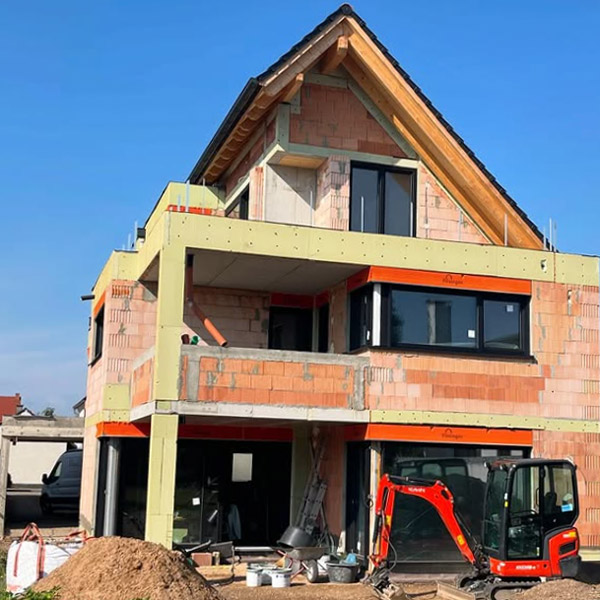 Neubau eines Hauses mit grauen Fenstern und rotem Dach. Bagger und Materialien im Vordergrund.