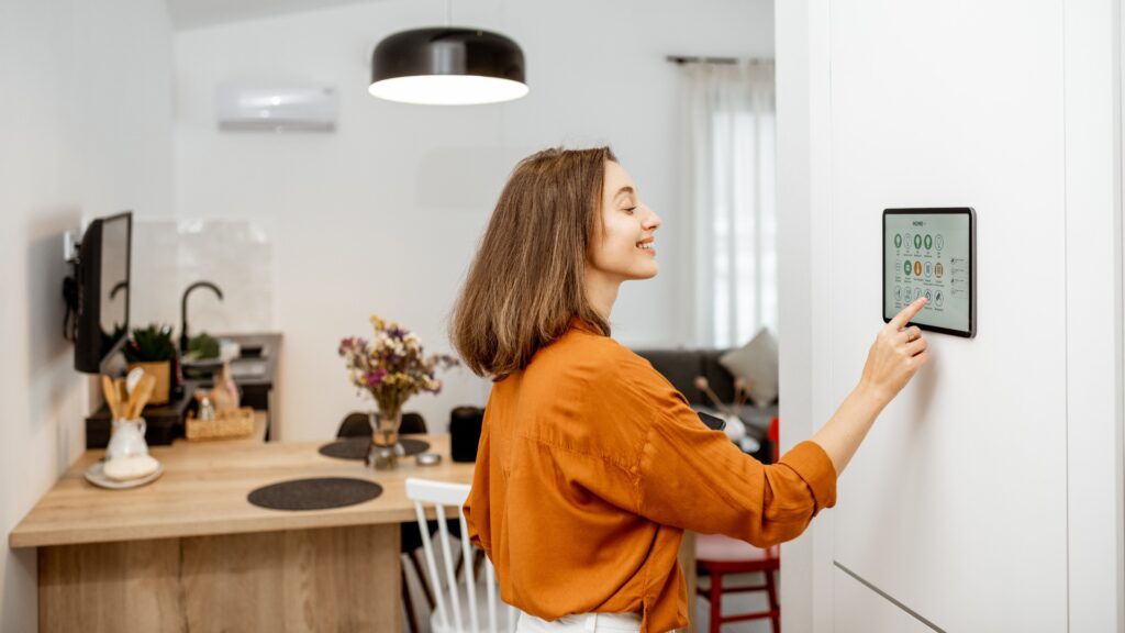 Junge Frau steuert ihr Zuhause über ein digitales Touchscreen-Panel, das an der Wand im Wohnzimmer installiert ist. Konzept eines Smart Homes und einer mobilen Anwendung zur Verwaltung von Smart-Geräten zu Hause.