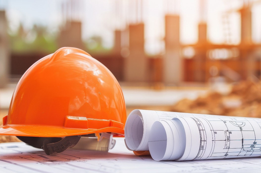 Nahaufnahme eines orangefarbenen Bauhelms und gerollter Baupläne, die auf einem Tisch auf einer Baustelle liegen und Planung und Baumanagement symbolisieren.